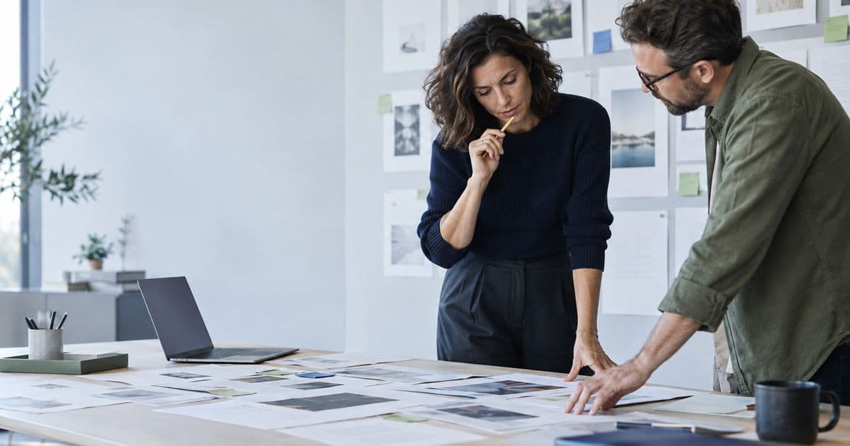 Two people review a spread of printouts and a wall of visual reference materials in a bright workroom.