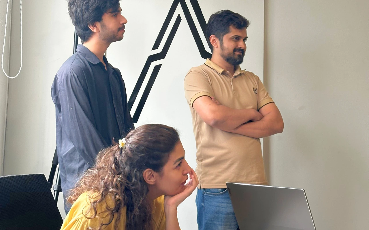 A female team member sits at a laptop while two male colleagues stand behind her, all concentrating on the screen in a modern office.