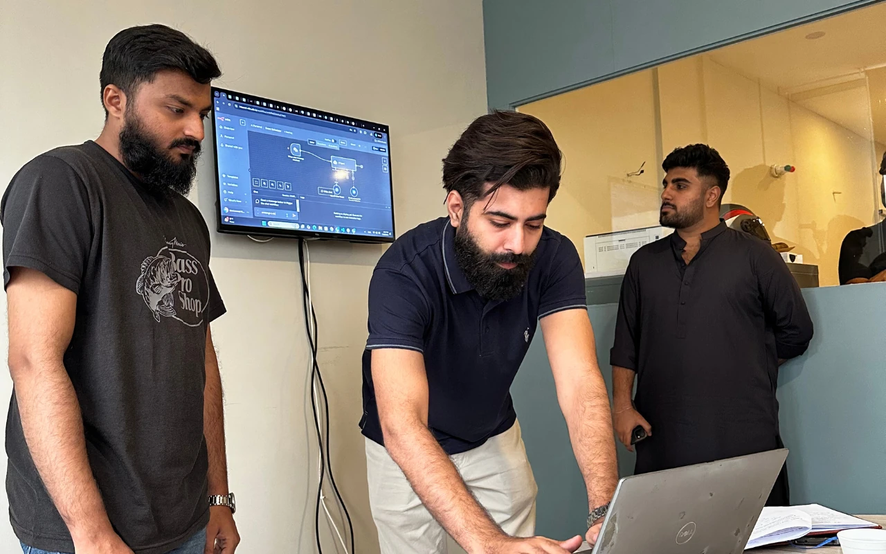 Three male engineers collaborating over a complex diagram shown on both a laptop and a wall-mounted monitor.