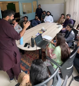 A full team meeting in an office conference room, viewed from a high angle. The team, including both men and women, is gathered around a long table with a laptop open, engaged in a discussion