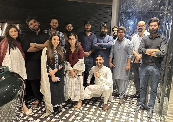 A diverse group of ten male and three female team members posing for a casual group photo indoors at night, showcasing a relaxed team gathering or social event.