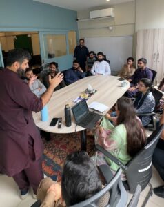 A full team meeting in an office conference room, viewed from a high angle. The team, including both men and women, is gathered around a long table with a laptop open, engaged in a discussion